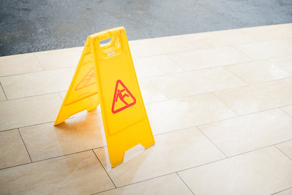 A yellow wet floor caution sign with red hazard symbols is placed on a light beige ceramic tiled floor in an indoor or outdoor area. The tiles are clean, smooth, and reflective under the lighting. The sign is positioned near the edge of the tiled surface, which transitions to a darker asphalt or concrete area outside. This surface cleaning and safety sign highlight the importance of maintaining dry, hazard-free flooring in residential or commercial settings, as part of professional cleaning and hygiene practices offered by [COMPANY_NAME], particularly relevant for end of tenancy and deep cleaning services on the Clapham cleaners.com page about avoiding hidden charges.
