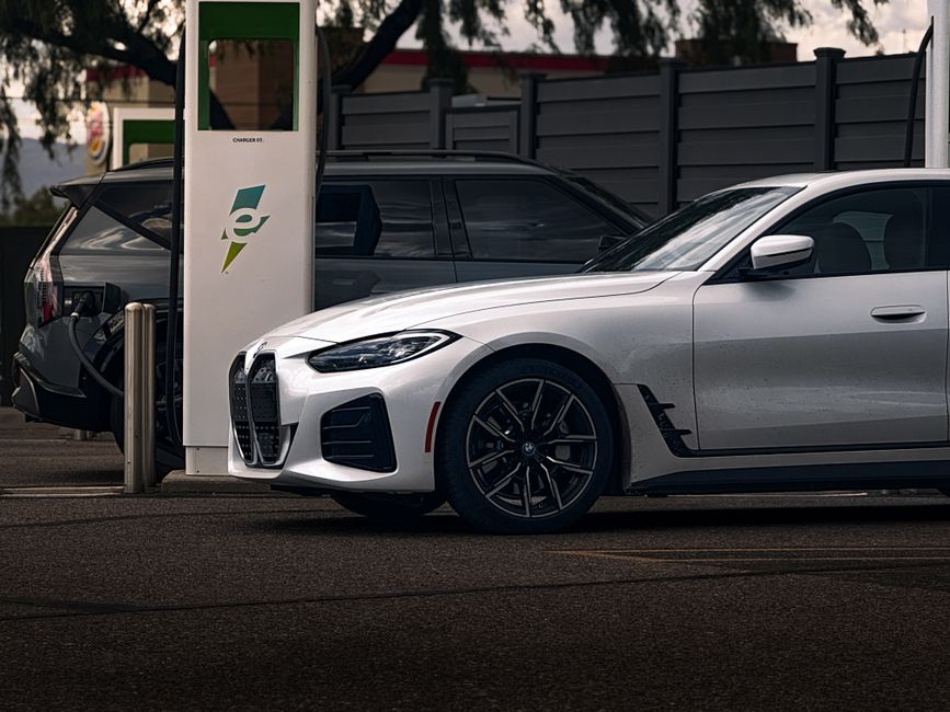 A white sports car parked at an electric vehicle charging station, with the front side prominently displayed, showing a sleek design, black alloy wheels, and smooth bodywork. The charging station is behind the car, featuring a green and white logo, and connected to a charging cable. Adjacent to the white car, a dark-colored SUV is also plugged into a separate charging point; both vehicles are situated on a paved surface under natural daylight. The background includes a grey wooden fence and trees, indicating an outdoor setting. This image reflects the process of electric vehicle charging in a clean, well-maintained parking area, associated with contemporary sustainable mobility, as seen on claphamcleaners.com for domestic and commercial surface cleaning references.