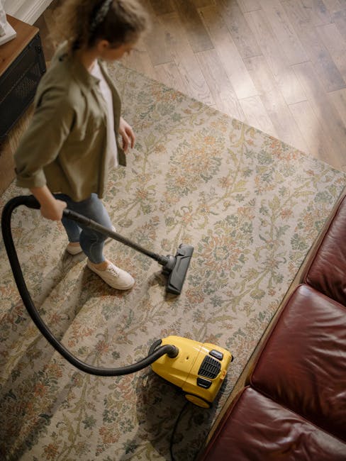 A professional cleaner dressed in a white protective suit, gloves, and mask is performing deep cleaning on a beige carpet in a living room. They are using a vacuum and steam extraction equipment, with visible steam or fog around the cleaner's feet, indicating the sanitisation process. The room features a modern, minimalist design with a wooden bookshelf filled with books and decorative items, a large potted plant, and natural daylight illuminating the space. The surface cleaning highlights cleanliness and hygiene, consistent with domestic cleaning services offered by Clapham Cleaners. The setting emphasizes effective carpet cleaning and sanitisation in a residential environment.