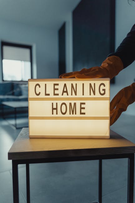 A close-up of a lightbox sign with illuminated letters reading 'CLEANING HOME' placed on a wooden surface, with a person wearing orange gloves reaching out behind the sign. The background features a modern, well-lit residential interior with large windows, blue-toned walls, and minimalistic furnishings. The scene emphasizes domestic cleaning and surface sanitisation, highlighting the importance of hygiene in home environments, as promoted by Clapham Cleaners. The clean and tidy setting reflects professional cleaning standards for residential spaces, suitable for the Clapham Common flat cleaning guide for landlords SW4.
