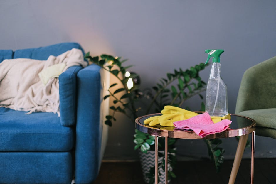 A living room scene featuring a blue upholstered sofa with a cream-colored blanket draped over one armrest, situated against a soft blue wall. Next to the sofa, there is a small round side table with a metallic finish, holding cleaning items including a clear spray bottle with a green nozzle, yellow rubber gloves, and a pink cleaning cloth. Behind the table, a green upholstered armchair is partially visible. In the background, a potted plant with broad green leaves adds a natural element to the room, and a modern wall-mounted light fixture provides subtle illumination. The surfaces, including the sofa fabric and side table, appear clean and well-maintained, reflecting effective domestic surface cleaning and hygiene practices consistent with the Clapham Common flat cleaning guide for landlords SW4, supported by Clapham cleaners.com.