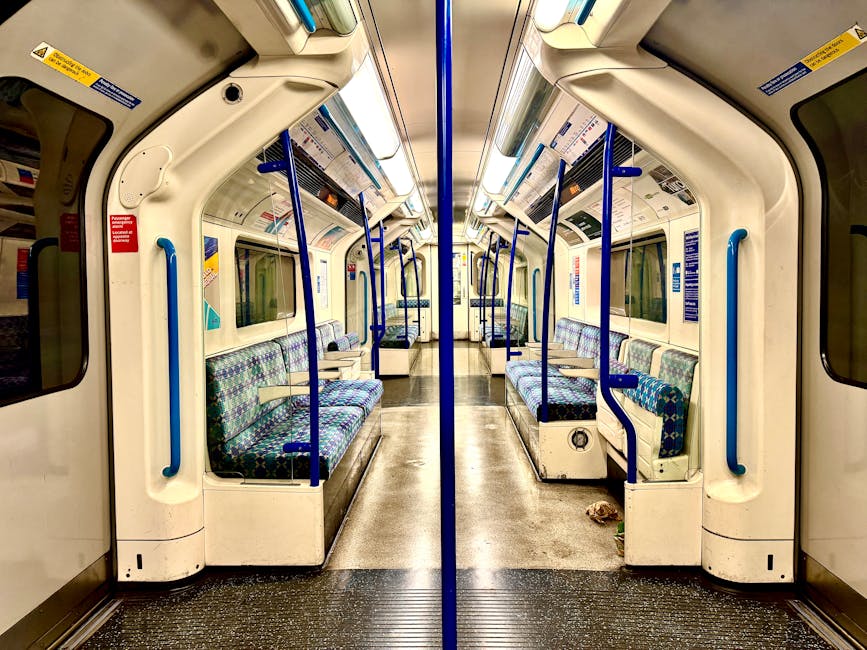 Interior of an empty train carriage near SW11 station, featuring a clean and well-maintained environment with patterned fabric seats in shades of blue, green, and purple. The seats are arranged in pairs along both sides of the carriage, with blue handrails and vertical support bars near the doors. The floor is made of dark textured material, appearing spotless and dry. Overhead, there are white storage compartments and lighting fixtures, creating a bright atmosphere. The large windows on either side show reflections of the interior, and the carriage's surfaces appear freshly cleaned, emphasizing hygiene and sanitation. This setting highlights the importance of surface cleaning and maintenance within public transport, as promoted by Clapham cleaners.com, to ensure hygienic and pleasant environments for commuters.