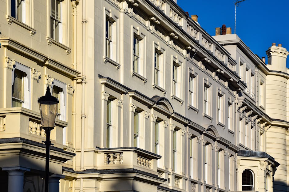 The image shows a close-up of a classic Victorian-style building with ornate architectural details, including decorative cornices, window trims, and small balconies with balustrades. The façade is painted in a pale cream color, reflecting sunlight, and features multiple large, rectangular windows with white frames and sills. A traditional black street lamp is positioned in the foreground, standing upright against the building, which is situated on a street with clear blue skies above. The overall scene captures the exterior of a well-maintained, historic residential building, emphasizing its detailed masonry and bright, clean appearance. This setting highlights the importance of surface cleaning, maintenance, and preservative care, possibly related to professional cleaning services offered by claphamcleaners.com, such as deep cleaning or building sanitisation in residential or commercial properties.