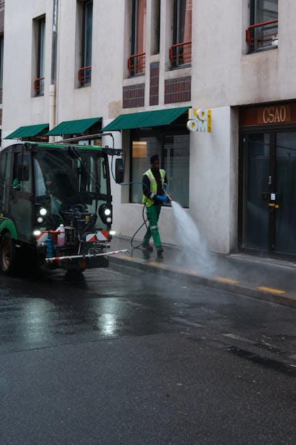 A worker wearing a high-visibility vest and gloves is operating a street cleaning machine that sprays water and cleans the pavement in front of a multi-storey commercial building with a white facade, large windows, and green awnings. The scene is set outdoors during daylight hours, with water pooling on the black asphalt surface, indicating a deep cleaning process as part of a professional sanitisation or surface cleaning service. The building's entrance features a glass door and is adjacent to signage, suggesting a commercial or office space, with the overall area appearing well-maintained and freshly cleaned. The presence of the cleaning machine and the worker highlights thorough cleaning and sanitation efforts typical for end of tenancy or deep cleaning services provided by Clapham Cleaners.