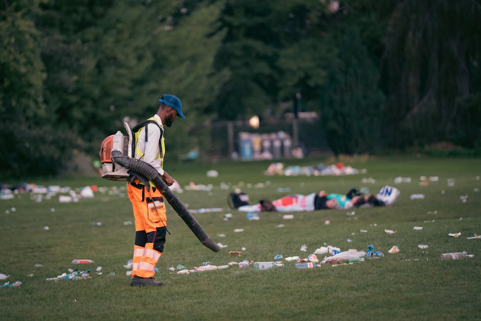 A man wearing high-visibility orange trousers, a blue cap, and a white jacket is standing on a grassy park area, using a leaf blower to clear away scattered litter and debris from the ground. The surface is natural grass, visibly strewn with plastic bottles, paper, and other waste, indicating a recent outdoor event or gathering. In the background, several individuals are lying on the grass, possibly resting or sleeping, with trees and a small shed or structure further behind. The scene is illuminated by natural lighting, highlighting the contrast between the cleanliness efforts by [COMPANY_NAME] and the surrounding litter, emphasizing the importance of surface cleaning and waste management in maintaining outdoor hygiene.