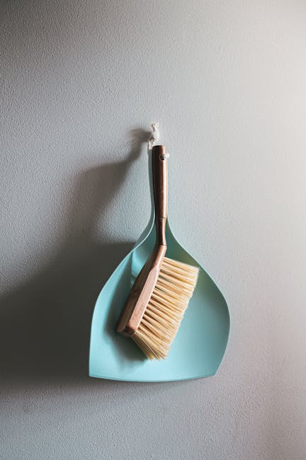 A close-up of a light grey wall with a wall hook holding a handheld dustpan and brush set, used in domestic surface cleaning. The dustpan is made of pale blue plastic with a smooth surface and is hanging flat against the wall. Resting inside the dustpan is a natural wooden-handled broom with creamy bristles, positioned vertically. The background shows a clean, minimally decorated section of a room, with even lighting highlighting the neatness and readiness for cleaning tasks, illustrating the type of tools used for everyday home cleaning and maintenance. This setup is representative of a typical cleaning supply storage area in residential spaces, emphasizing hygiene and surface cleanliness, relevant to same day cleaning delays in Clapham common problems and fixes.